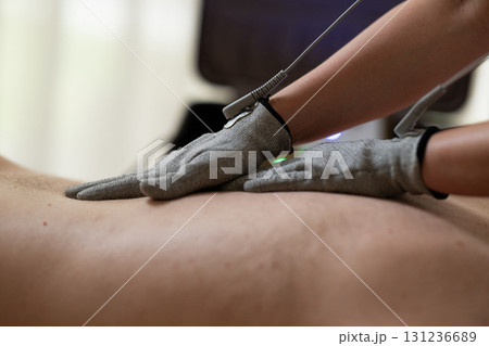 Close up view of a therapist applying electrotherapy with special conductive gloves on a man back during a physiotherapy session. Concept of modern rehabilitation, pain relief, muscle stimulation, and Close up view of a therapist applying electrotherapy with special conductive gloves on a man back during a physiotherapy session. Concept of modern rehabilitation, pain relief, muscle stimulation, and 131236689