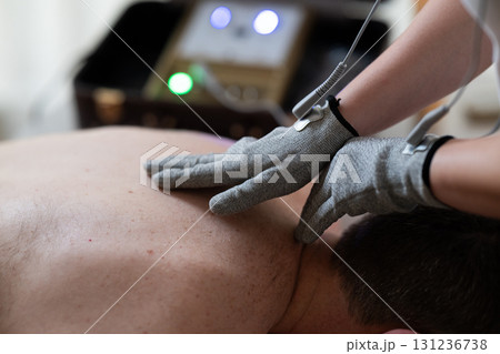 Close up view of a therapist applying electrotherapy with special conductive gloves on a man back during a physiotherapy session. Concept of modern rehabilitation, pain relief, muscle stimulation, and Close up view of a therapist applying electrotherapy with special conductive gloves on a man back during a physiotherapy session. Concept of modern rehabilitation, pain relief, muscle stimulation, and 131236738