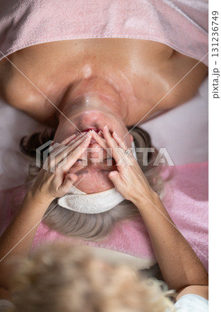 Close up of a mature woman lying on a treatment bed with a towel and headband while a professional therapist applies foam during a facial cleansing procedure. Skincare and wellness concept Close up of a mature woman lying on a treatment bed with a towel and headband while a professional therapist applies foam during a facial cleansing procedure. Skincare and wellness concept 131236749