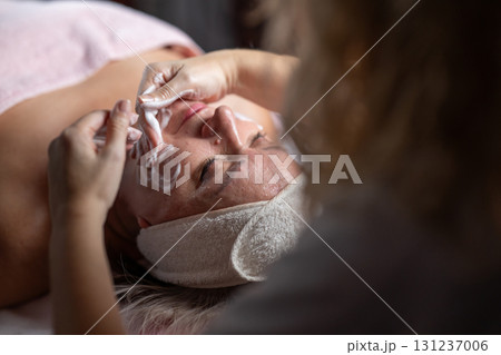 Close up of a mature woman lying on a treatment bed with a towel and headband while a professional therapist applies foam during a facial cleansing procedure. Skincare and wellness concept 131237006