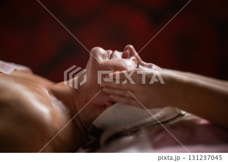 Side view close up of a mature woman lying on a treatment bed with a headband while a professional therapist massages her face. 131237045