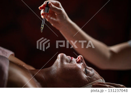Close up of a mature woman lying in a spa while a therapist applies skincare serum with a dropper above her face. Beauty and wellness concept highlighting anti aging treatment, hydration, and holistic Close up of a mature woman lying in a spa while a therapist applies skincare serum with a dropper above her face. Beauty and wellness concept highlighting anti aging treatment, hydration, and holistic 131237048