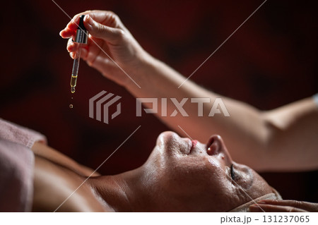 Close up of a mature woman lying in a spa while a therapist applies skincare serum with a dropper above her face. Beauty and wellness concept highlighting anti aging treatment, hydration, and holistic 131237065