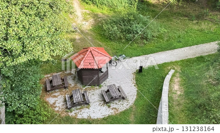 A wooden gazebo with a red roof is surrounded by green grass and trees. Nearby, there are picnic tables and a pathway leading through the area 131238164