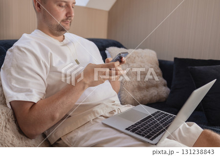 Man working on laptop and using smartphone while sitting on sofa at home. Concept of freelancing, digital lifestyle, mobile technology, e-banking, online shopping, communication and productivity.  131238843