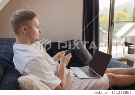Man working on laptop and using smartphone while sitting on sofa at home. Concept of freelancing, digital lifestyle, mobile technology, e-banking, online shopping, communication and productivity. Man working on laptop and using smartphone while sitting on sofa at home. Concept of freelancing, digital lifestyle, mobile technology, e-banking, online shopping, communication and productivity. 131238865