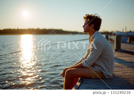 A man sits on a dock by a body of water, looking out at the sun 131240216