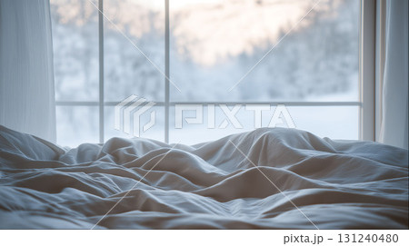 Messy cozy bed with rumpled sheets near large window overlooking peaceful snowy winter landscape 131240480