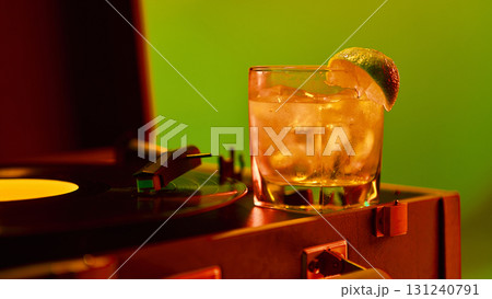 Cocktail with lime slice on reflective surface near record player under neon green background 131240791