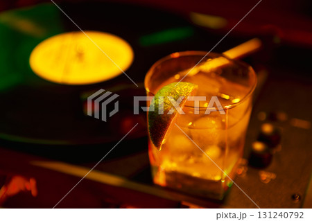 Close up of cocktail with lime wedge and ice cubes beside spinning vinyl in neon lighting Close up of cocktail with lime wedge and ice cubes beside spinning vinyl in neon lighting 131240792