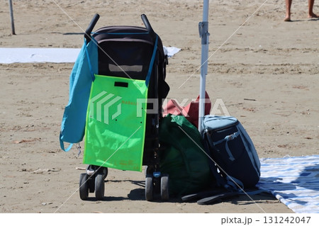 Baby stroller and bags parked on sandy beach under beach umbrella Baby stroller and bags parked on sandy beach under beach umbrella 131242047