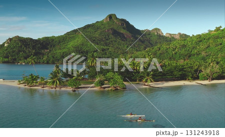 Two people paddling an outrigger canoe near tropical island with white sand beach, green towering mountains in background. Active sports lifestyle. French Polynesia. remote wild nature paradise aerial 131243918