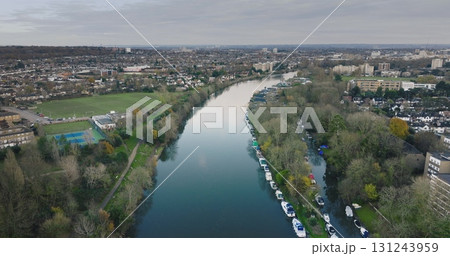 London city , Teddington: River Thames winding through Teddington, showcasing moored boats, residential houses, and green spaces on a cloudy day. UK capital cityscape. Aerial view drone flight 131243959