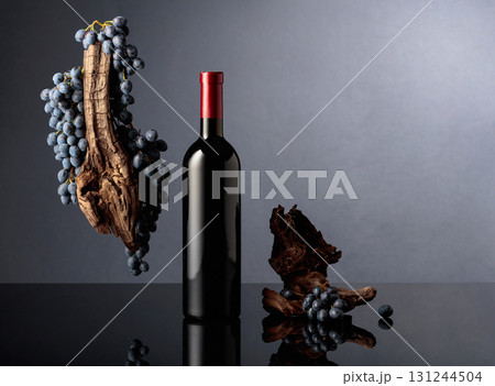 Bottle of red wine with a weathered snag and blue grapes on a black reflective background. 131244504