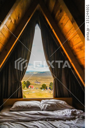 Cozy rustic bedroom in a wooden A-frame cabin with white pillows and blanket, window view of hills and open field in Montenegro. Cozy rustic bedroom in a wooden A-frame cabin with white pillows and blanket, window view of hills and open field in Montenegro. 131244801