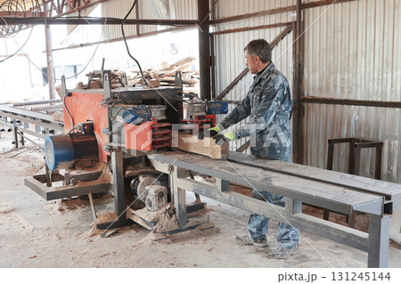 Male worker stands at woodworking machine. Woodworking company. Process of processing logs at sawmill. Sawing tree trunk. Sawmill. Male worker stands at woodworking machine. Woodworking company. Process of processing logs at sawmill. Sawing tree trunk. Sawmill. 131245144