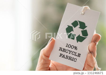Hand holding recycled fabric tag on sustainable clothing stack, eco textile concept Hand holding recycled fabric tag on sustainable clothing stack, eco textile concept 131246671