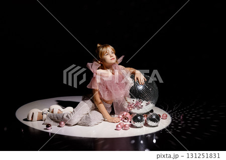Girl in pink top and sequin pants sitting on stage with disco balls and dark background 131251831