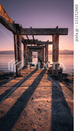 Old decaying wooden pier at sunset with long shadows and calm water, tranquil atmosphere Old decaying wooden pier at sunset with long shadows and calm water, tranquil atmosphere 131252660