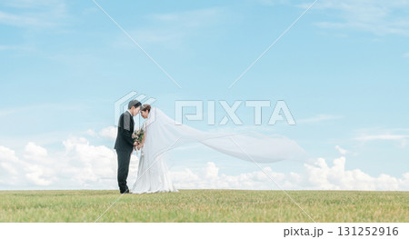 ベールをなびかせる空バックの新郎新婦（ウェディング・結婚式・ブライダル・フォトウェディング） 131252916