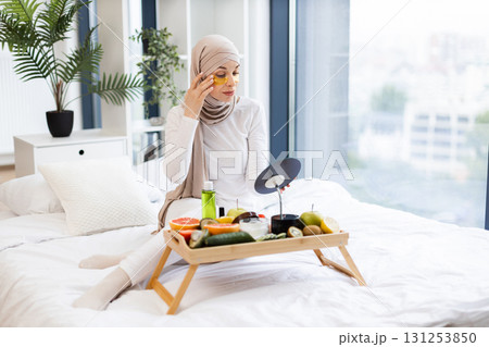 Young Muslim woman in beige hijab on bed handling beauty routine with hydrogel patches, surrounded by fresh fruits, skincare products. Image reflects self-care, health, lifestyle in cozy indoor. 131253850
