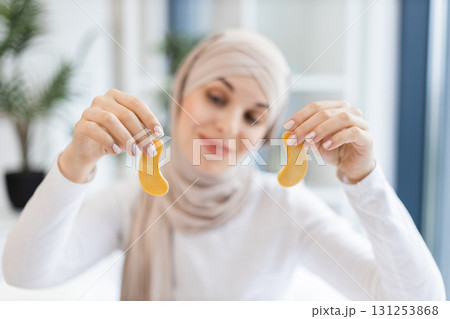Young Muslim woman wearing hijab holding skincare gold eye patches. Bright indoor setting with focus on self-care routine. Promoting relaxation, beauty treatment. 131253868