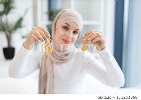 Young Muslim woman wearing hijab holding skincare gold eye patches. Bright indoor setting with focus on self-care routine. Promoting relaxation, beauty treatment. 131253869
