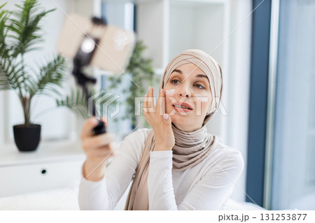 Young adult Muslim woman with hijab applying facial cream while using smartphone for vlogging. Captures concept of modern self-care and engagement through social media. 131253877