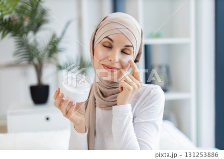 Young adult Muslim woman in hijab skincare routine at home with facial cream. Expresses happiness and simple beauty self-care. Bright indoor atmosphere showcasing plant in stylish decor Young adult Muslim woman in hijab skincare routine at home with facial cream. Expresses happiness and simple beauty self-care. Bright indoor atmosphere showcasing plant in stylish decor 131253886