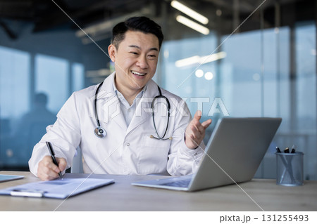 Asian young male doctor consulting online, sitting in hospital office at laptop, writing in notebook and talking remotely. Asian young male doctor consulting online, sitting in hospital office at laptop, writing in notebook and talking remotely. 131255493