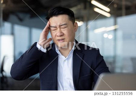 Asian young male businessman sitting in the office at his desk and holding his head with his hand feeling severe pain and pressure. Asian young male businessman sitting in the office at his desk and holding his head with his hand feeling severe pain and pressure. 131255556
