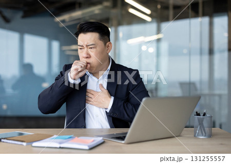 Sick young Asian man sitting in the office at his desk, closing his mouth and coughing, holding his hand to his chest. Sick young Asian man sitting in the office at his desk, closing his mouth and coughing, holding his hand to his chest. 131255557
