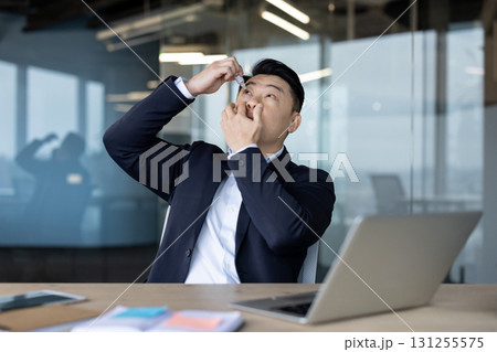 Tired Asian businessman sitting at work and working overtime, applying eye drops to relieve pain and tension. Tired Asian businessman sitting at work and working overtime, applying eye drops to relieve pain and tension. 131255575