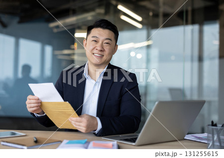 Portrait of a smiling young Asian man in a suit sitting in the office at the desk, holding an open envelope with a letter in his hands and looking at the camera. 131255615