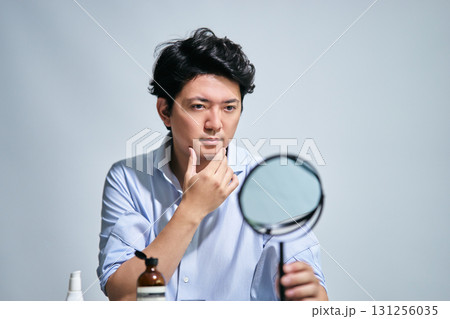 メンズコスメの道具を並べた机で鏡を見つめてスキンケアするシャツを着た30代の男性 メンズコスメの道具を並べた机で鏡を見つめてスキンケアするシャツを着た30代の男性 131256035