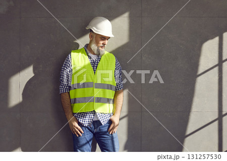 Portrait of mature bearded foreman in a helmet and construction vest on grey wall background. 131257530