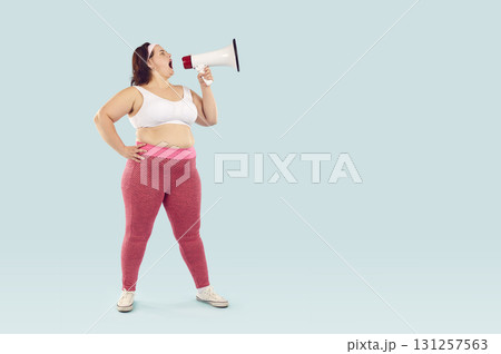 Young funny fat woman in sportswear screaming with mouthpiece on studio blue background. 131257563
