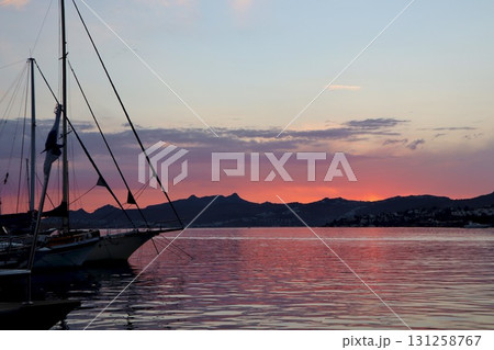 A serene coastal scene at sunset, with the sky and water glowing in hues of red and orange, as silhouettes of sailboats and distant hills are cast against the light. 131258767