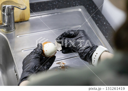 Hands in black protective gloves meticulously peel a freshly hard-boiled chicken egg over a clean stainless steel kitchen sink, ensuring safe food preparation Hands in black protective gloves meticulously peel a freshly hard-boiled chicken egg over a clean stainless steel kitchen sink, ensuring safe food preparation 131261419