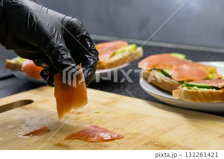 Chef's gloved hand expertly arranges fresh salmon fillet on a wooden board, crafting gourmet sandwiches, highlighting healthy food preparation and culinary skill Chef's gloved hand expertly arranges fresh salmon fillet on a wooden board, crafting gourmet sandwiches, highlighting healthy food preparation and culinary skill 131261421