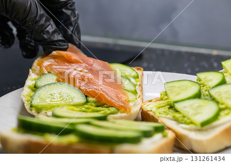 Gloved hand placing smoked salmon on a fresh avocado and cream cheese toast with cucumber slices, perfect for a healthy breakfast Gloved hand placing smoked salmon on a fresh avocado and cream cheese toast with cucumber slices, perfect for a healthy breakfast 131261434