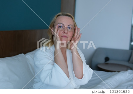 woman applying sheet mask at home in front of double sided mirror.beautiful girl in white robe sitting with coffee or tea cup in hand. relax and wellbeing moment. lot of cosmetic in front of female. woman applying sheet mask at home in front of double sided mirror.beautiful girl in white robe sitting with coffee or tea cup in hand. relax and wellbeing moment. lot of cosmetic in front of female. 131261626