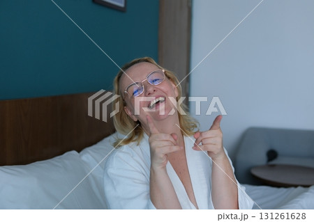 woman applying sheet mask at home in front of double sided mirror.beautiful girl in white robe sitting with coffee or tea cup in hand. relax and wellbeing moment. lot of cosmetic in front of female. woman applying sheet mask at home in front of double sided mirror.beautiful girl in white robe sitting with coffee or tea cup in hand. relax and wellbeing moment. lot of cosmetic in front of female. 131261628