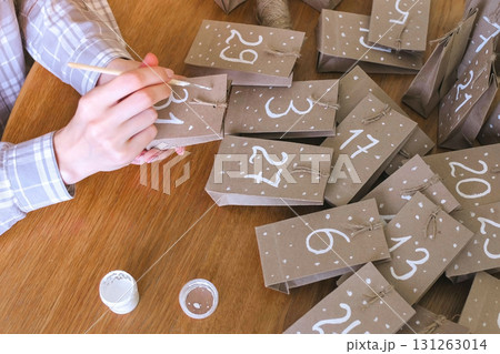 Woman makes christmas advent calendar for kids. Writes a number on the bag by brush and white paint. Close-up hands. 131263014