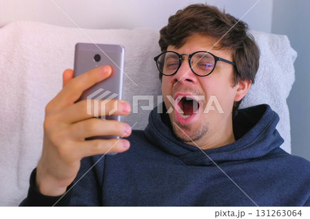 Sleepy man in glasses yawns and browsing in social media on smartphone sitting in armchair. 131263064