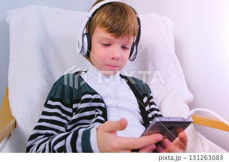 Boy is watching a video in headphones in mobile phone sitting in armchair. Boy is watching a video in headphones in mobile phone sitting in armchair. 131263083
