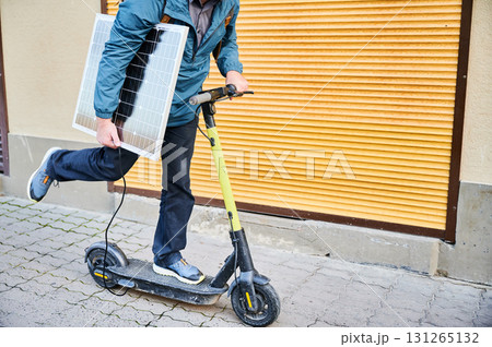 Cropped view of man rides electric scooter while carrying solar panel. Integration of solar power as sustainable energy source for charging electric scooters, promoting eco-friendly transportation. 131265132