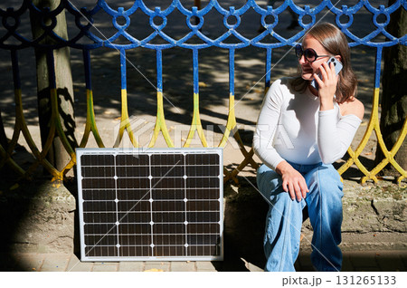 Happy woman using smartphone next to photovoltaic solar panel. Integration of sustainable renewable energy into everyday life, demonstrating practical use of solar power for charging devices. Happy woman using smartphone next to photovoltaic solar panel. Integration of sustainable renewable energy into everyday life, demonstrating practical use of solar power for charging devices. 131265133