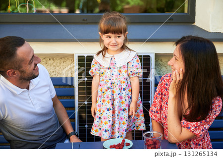 Happy family spending time together at sunny day. Little girl smiling to her parents. Young couple and their kid sitting outside on background of solar panel. Happy family spending time together at sunny day. Little girl smiling to her parents. Young couple and their kid sitting outside on background of solar panel. 131265134