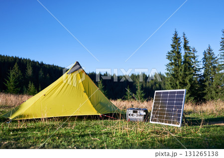 Tourist tent with solar panel and portable power station nearby in grassy field. Scene surrounded by rolling hills, tall pine trees under clear blue sky, showcasing eco-friendly camping setup. Tourist tent with solar panel and portable power station nearby in grassy field. Scene surrounded by rolling hills, tall pine trees under clear blue sky, showcasing eco-friendly camping setup. 131265138